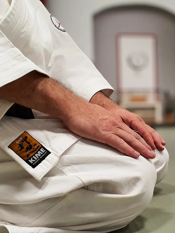 Seizában ülő aikidós keze a térdén, fehér aikidoruhában, a háttérben a dojo kamizája